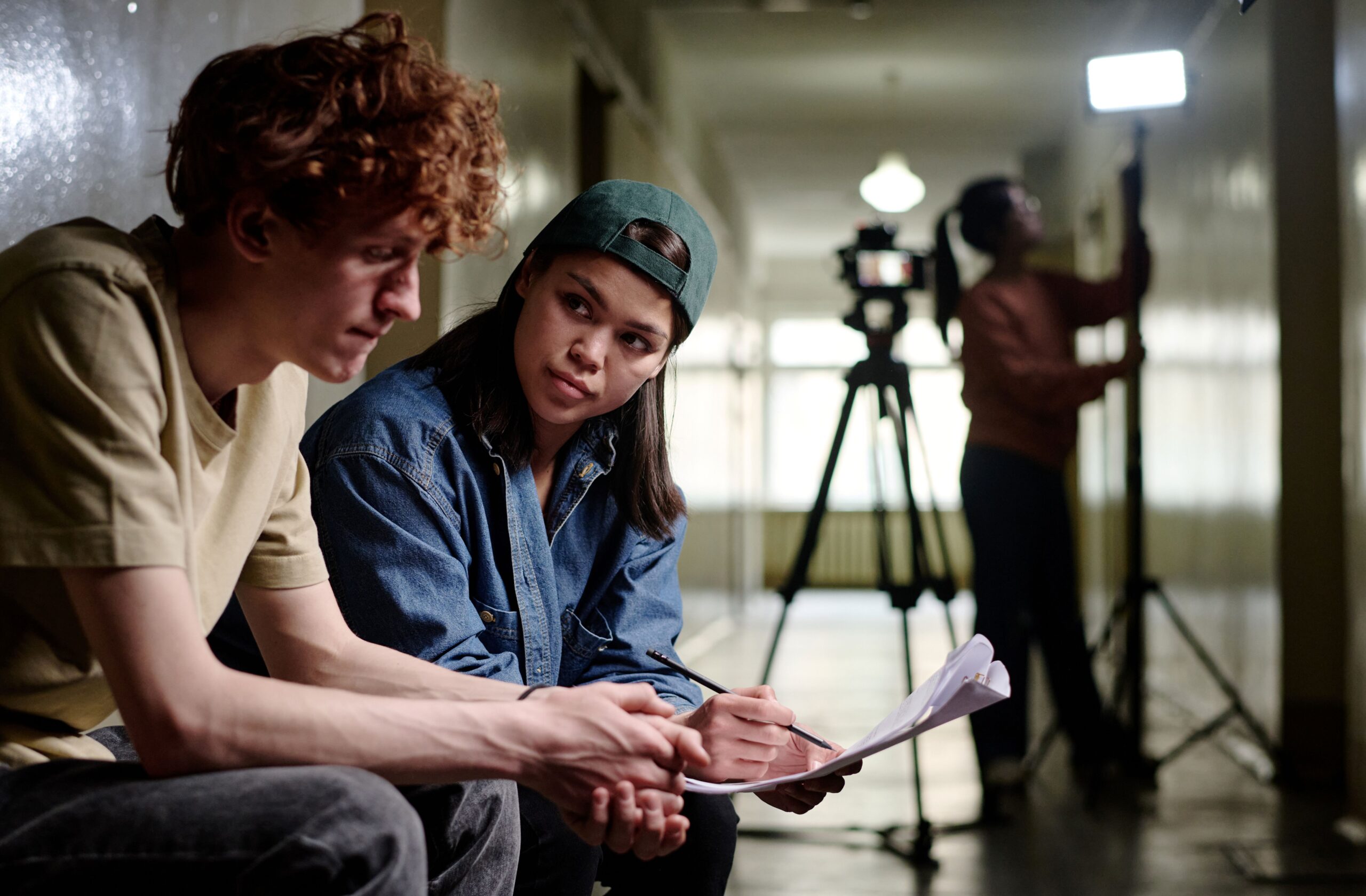 Filmproduktion hinter den Kulissen: Zwei Personen besprechen das Drehbuch, während im Hintergrund Kamera und Lichttechnik vorbereitet werden